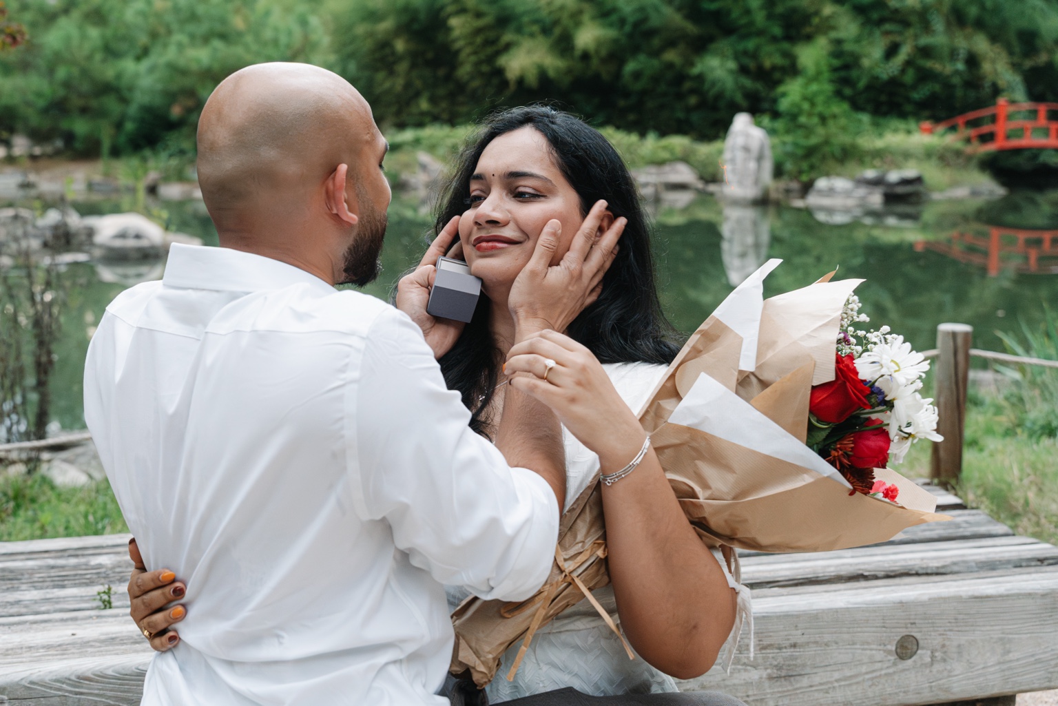 Akhil holding Maggi’s face as she holds flowers beside the pond with the red bridge behind them