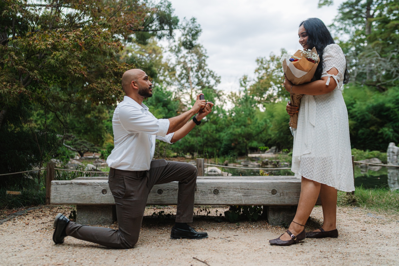 Akhil kneeling with an open ring box as Maggi smiles and holds a bouquet beside the garden pond