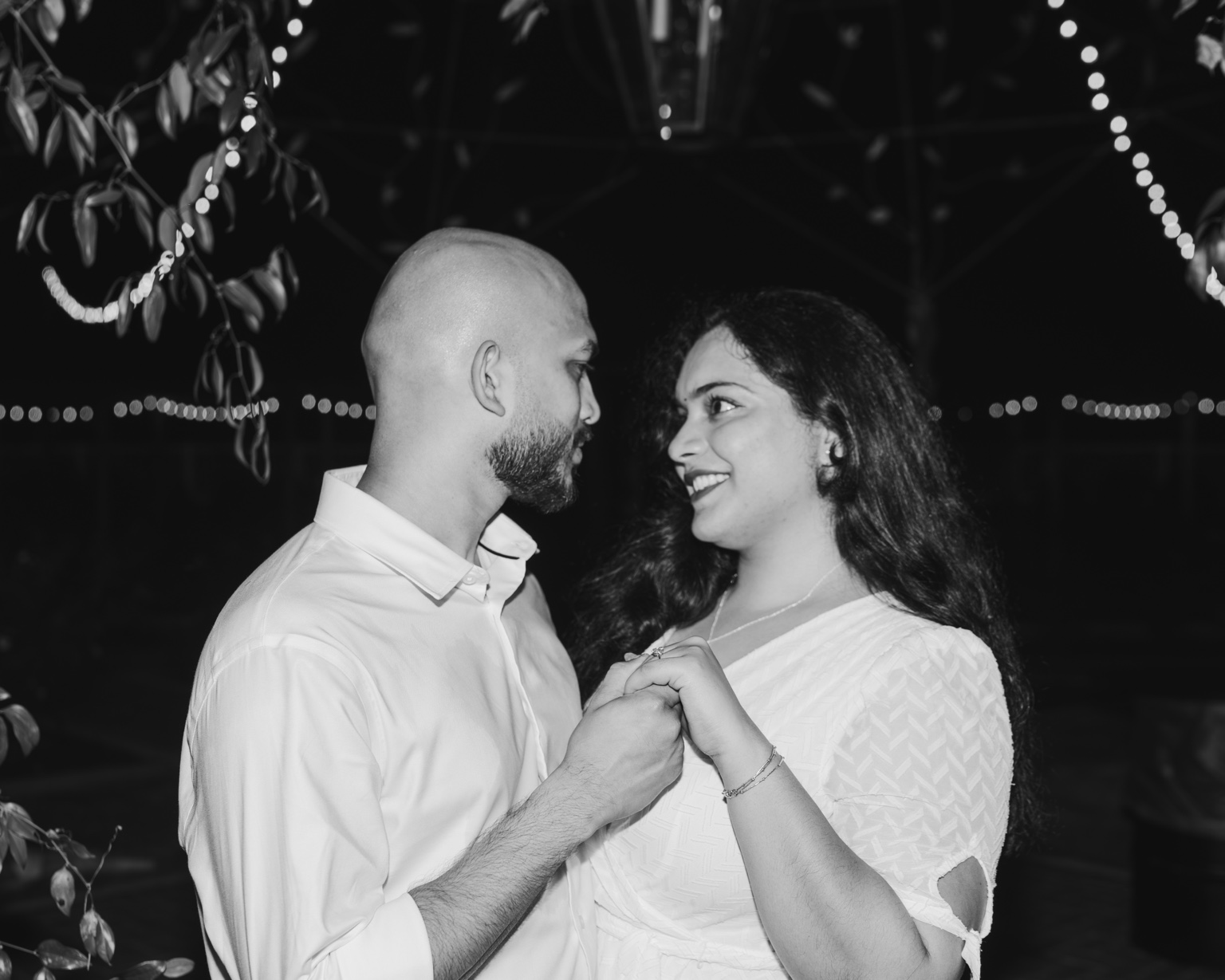 Black-and-white portrait of Akhil and Maggi holding hands and smiling at each other beneath string lights at night