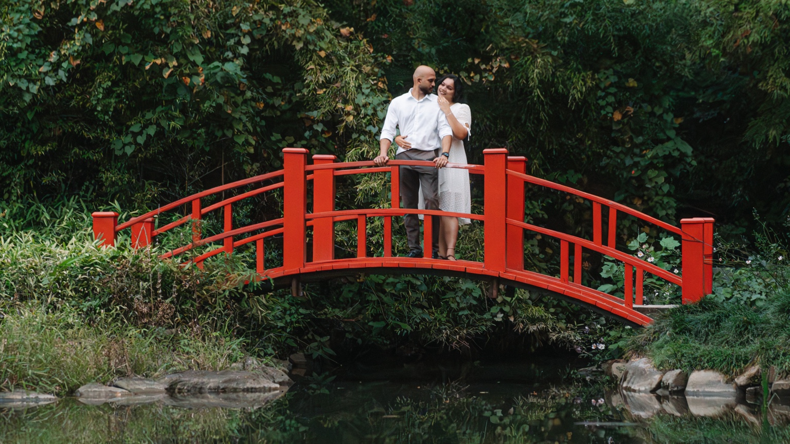 Akhil and Maggi embracing on the red bridge surrounded by green foliage at Birmingham Botanical Gardens
