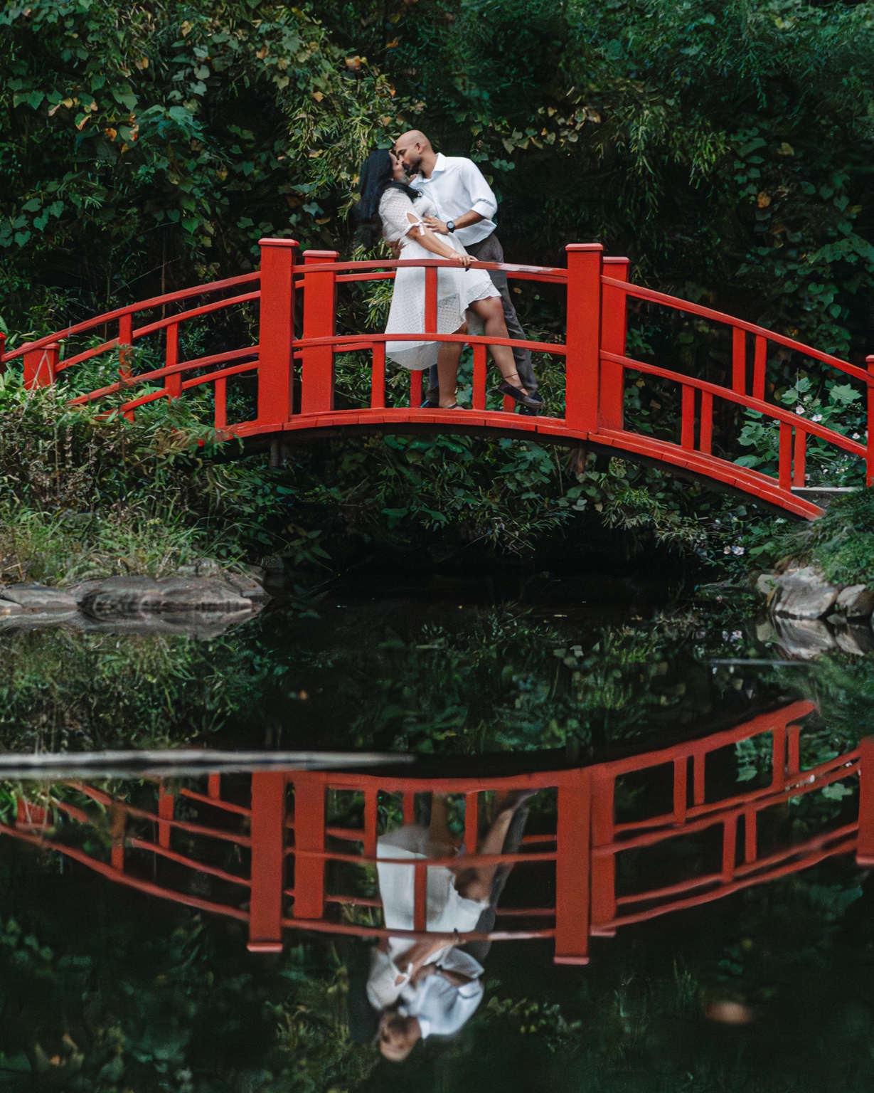Akhil and Maggi kissing on the red bridge with their reflection in the water at Birmingham Botanical Gardens